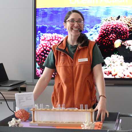 Presenter in orange vest sanding with chemistry set in front and picture of coral behind