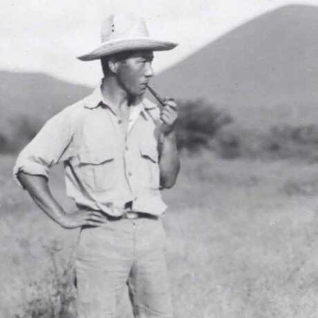 Toshio Asaeda stands in grasslands surrounded by mountains smoking a pipe