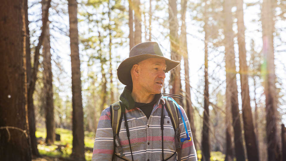Jack Dumbacher stands against a sunlit cluster of trees near Caples Creek, wearing a striped button down shirt and a wide brimmed hat. 